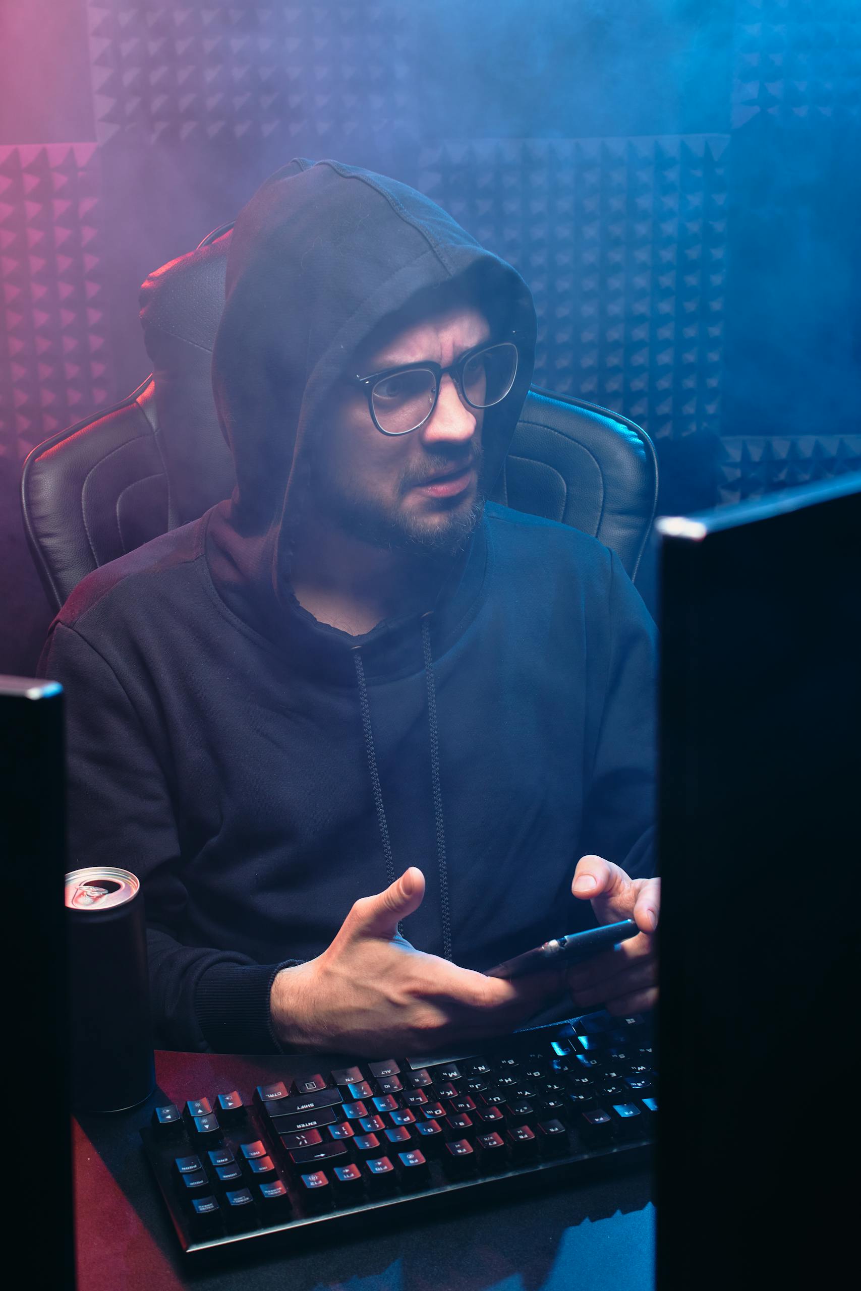 Man with hoodie using computer and smartphone in moody tech environment.